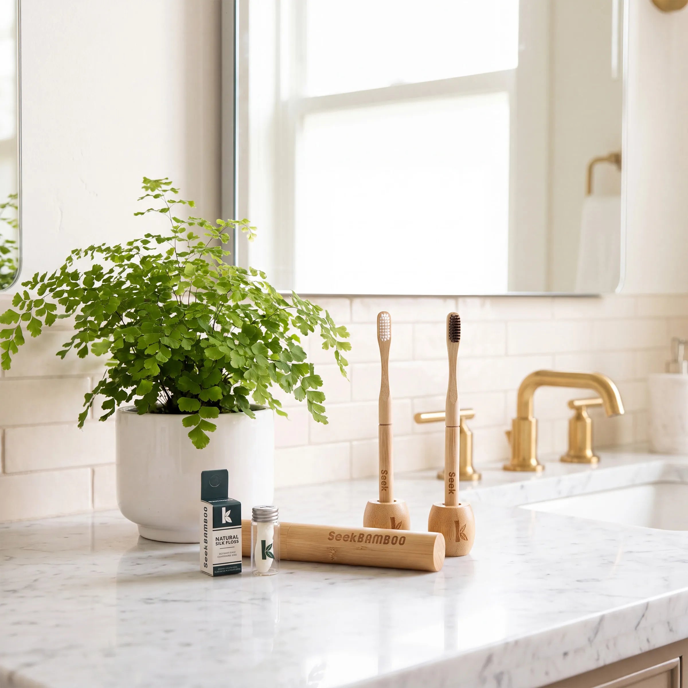 A bamboo toothbrush set displayed on a white marble bathroom counter, featuring two bamboo toothbrushes in wooden stands, a bamboo travel case, and a small branded box. A potted green fern and a gold faucet are visible in the background.