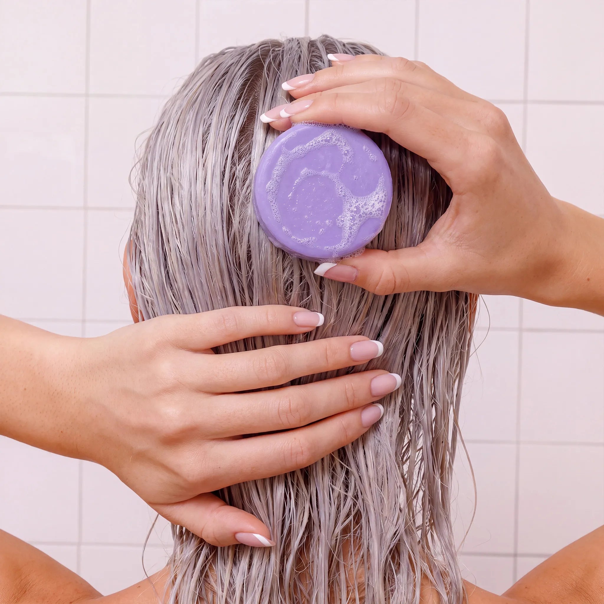 A person with wet, light purple-gray hair lathering it with a round lavender shampoo or conditioner bar, shown from the back against a pale tiled wall.