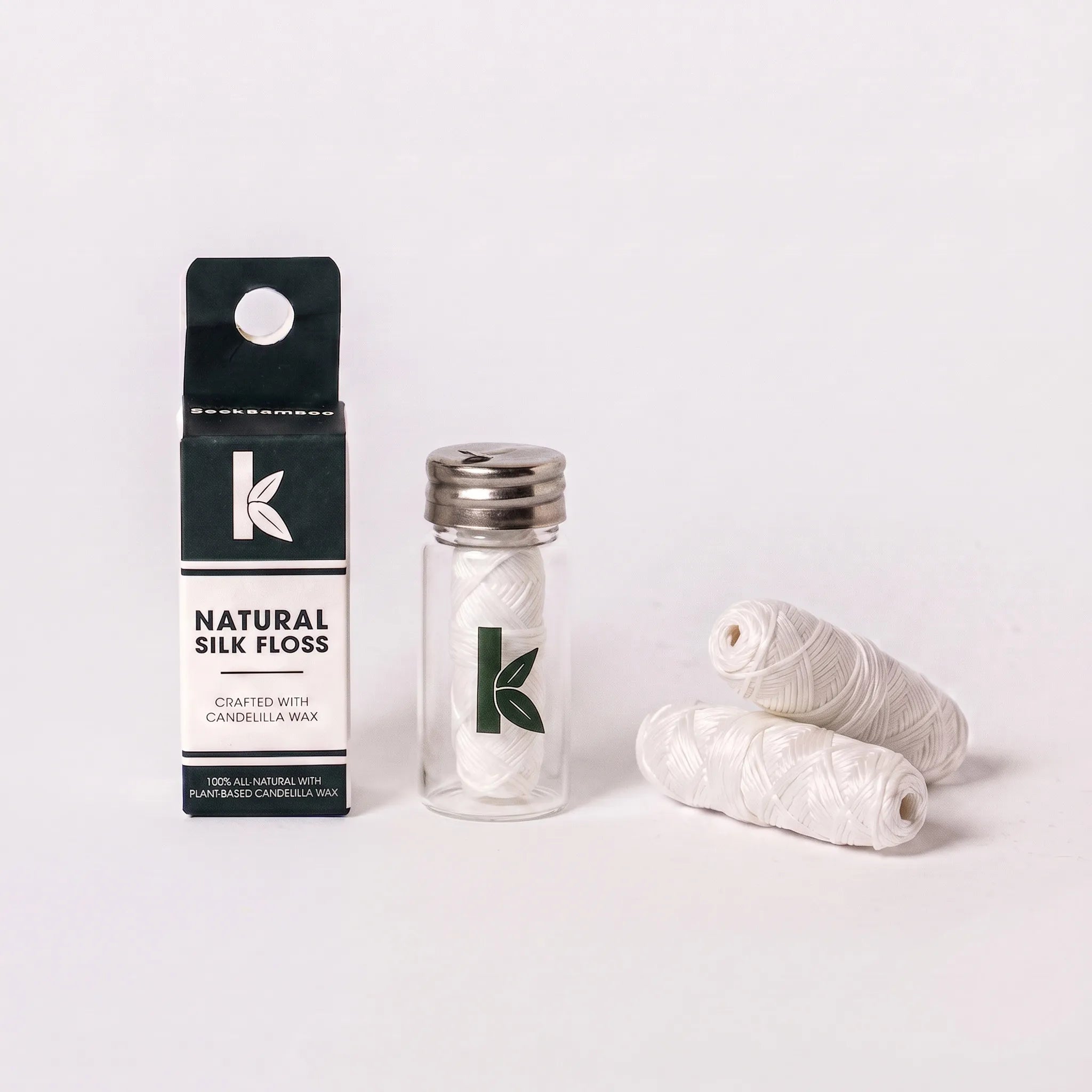 Natural silk floss shown with its black and white retail box, a clear glass jar with a silver lid, and three spools of white silk thread on a clean white background.