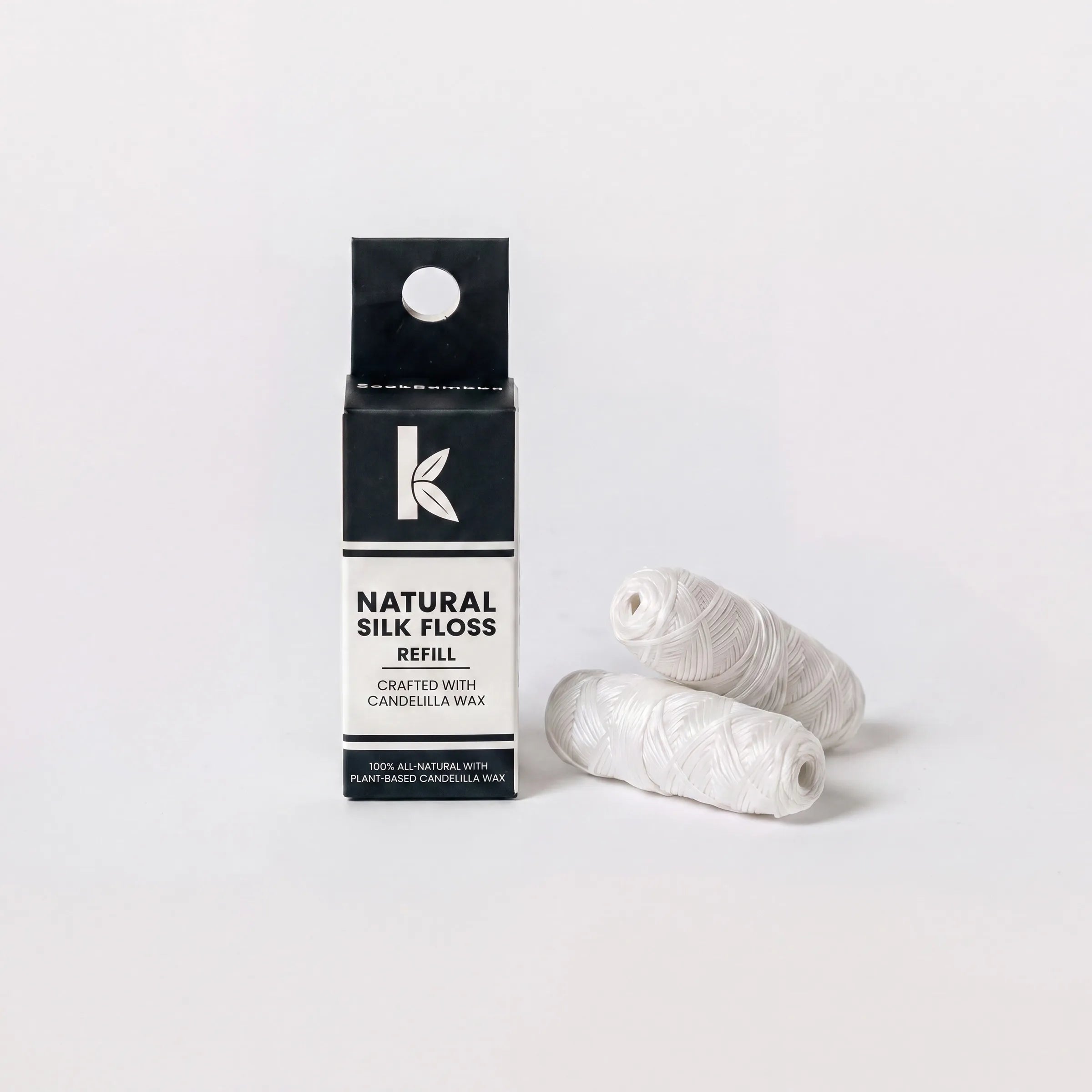 A black and white box of Natural Silk Floss Refill stands next to two white wound silk floss cones on a clean white background.