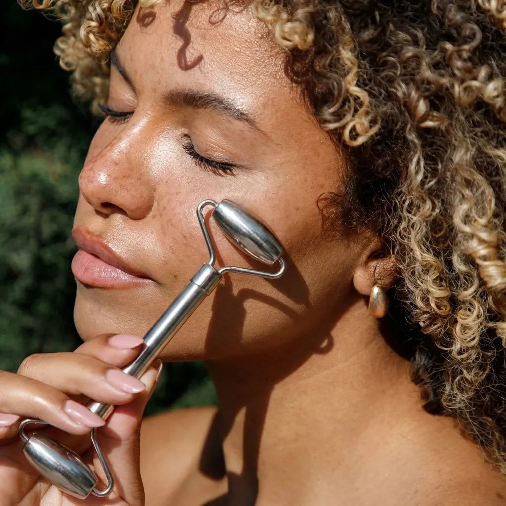 Woman using a stainless steel facial roller outdoors with greenery in the background
