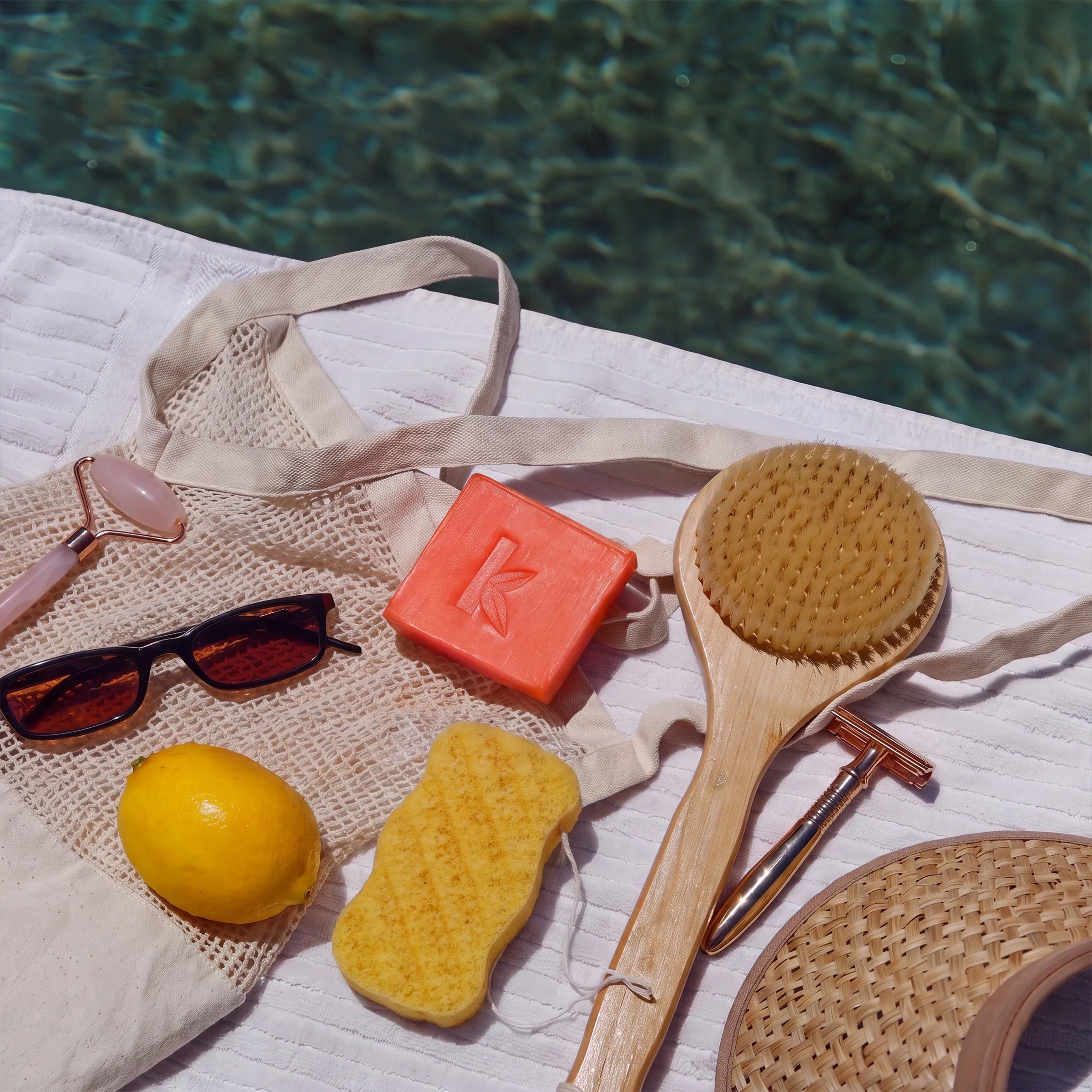 A styled flat-lay of spa and self-care items on a white towel beside a swimming pool: a natural wood shaving brush, a yellow sponge, an orange bar of soap, sunglasses, a lemon, a woven hat, a straw scoop, and a metal roller.