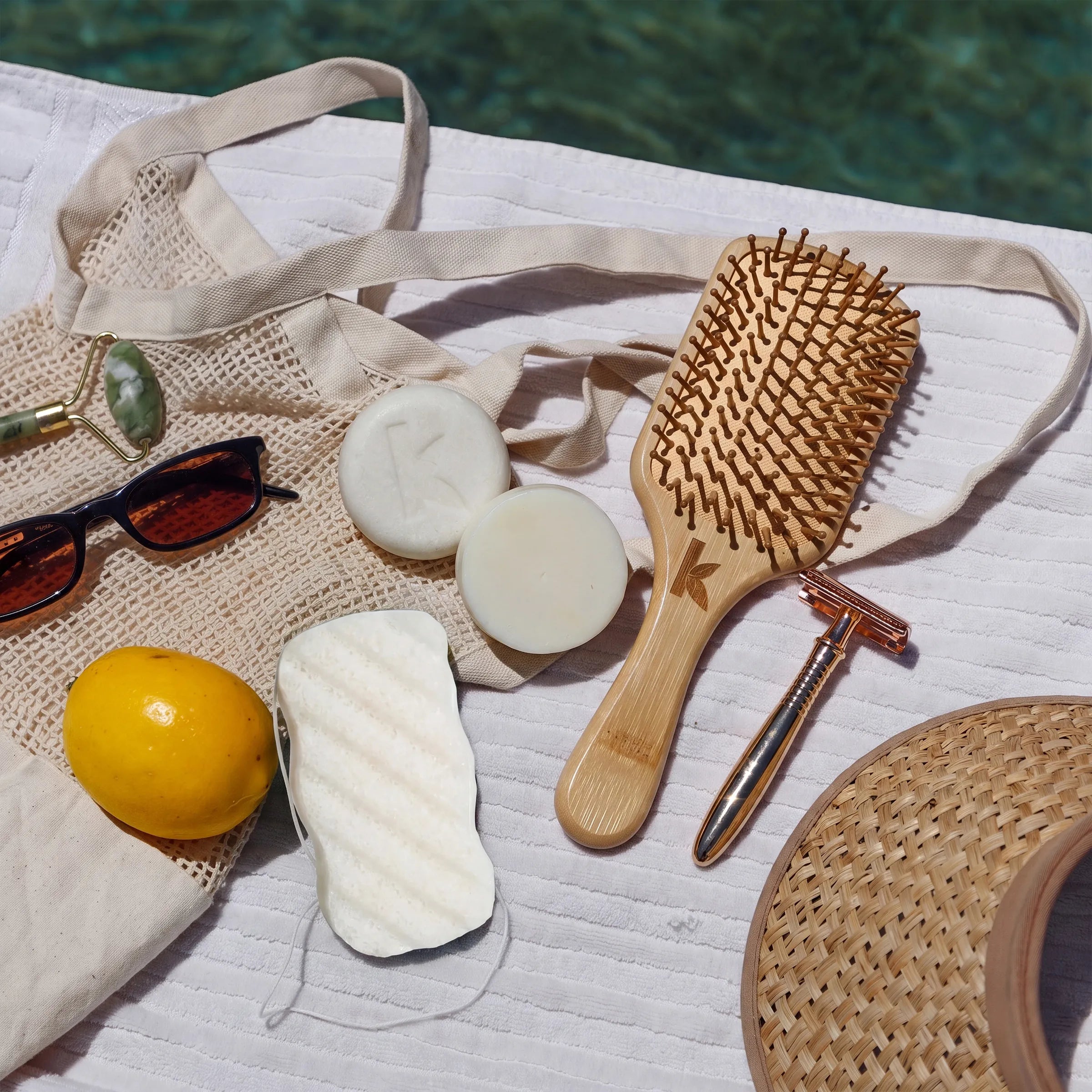 A styled flat-lay of beach and spa accessories on a white textured blanket near a pool, including a beige tote bag, wooden hairbrush, white soap bars, sunglasses, a yellow lemon, a metal razor, a woven hat edge, and a white body konjac sponge.