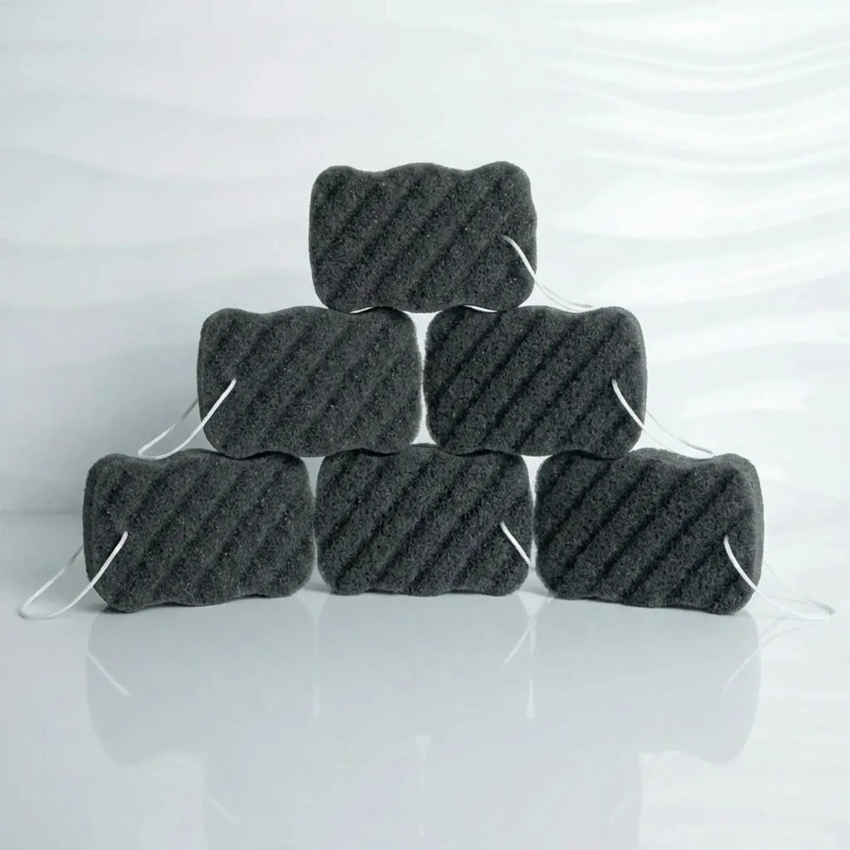 Six charcoal konjac body sponges with white elastic ties, stacked in a pyramid arrangement on a glossy white surface with a soft reflection.