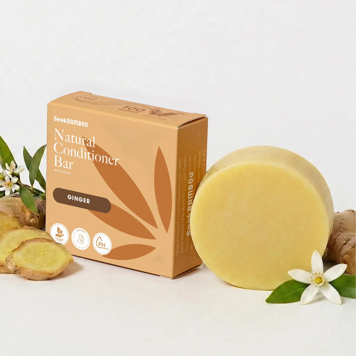 A round bar of a clarifying ginger conditioner bar next to its kraft paper packaging box, styled with ginger slices, a white flower, and green leaves on a clean white background.