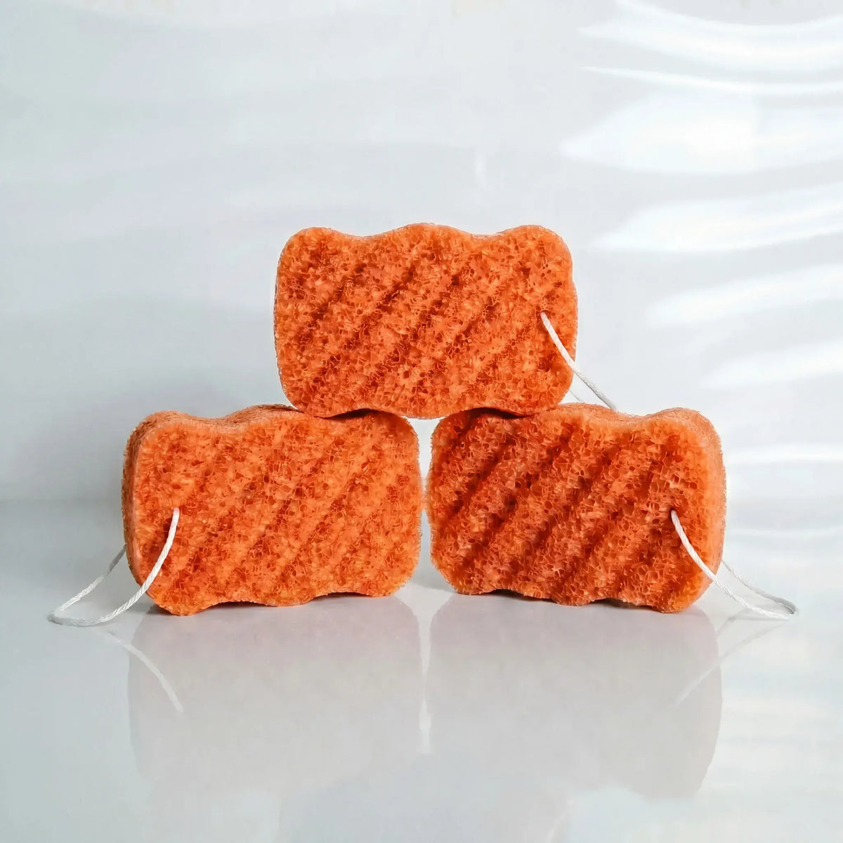 Three orange kojic acid konjac sponges stacked on a reflective surface with a white background