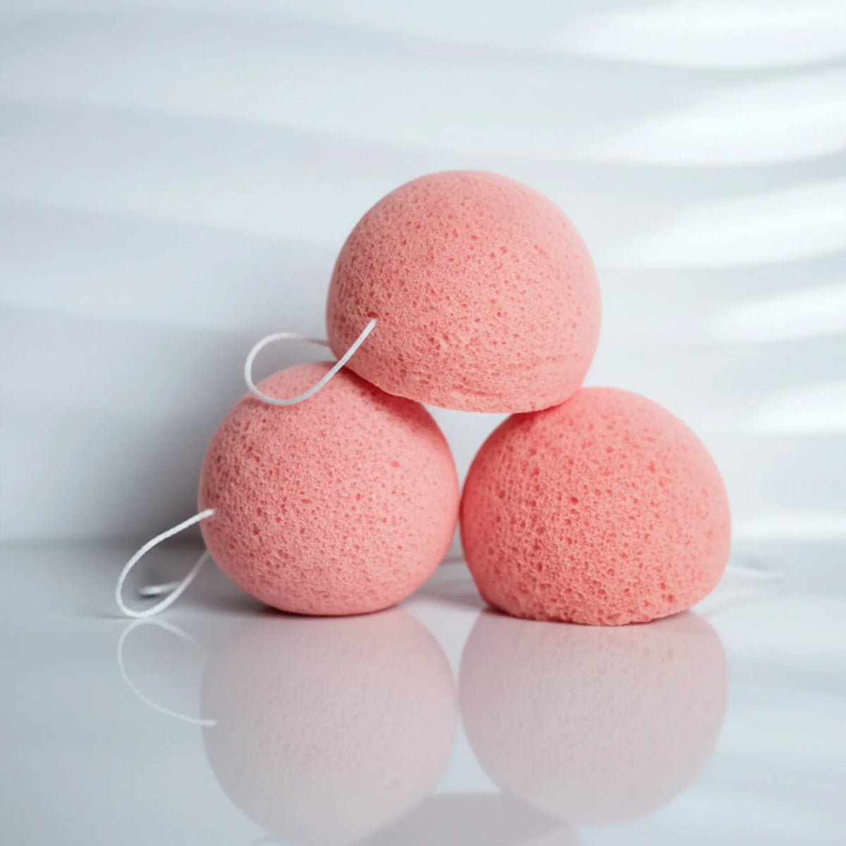 Three pink clay konjac sponges with white hanging loops, arranged in a triangular stack on a glossy white surface with soft shadows.
