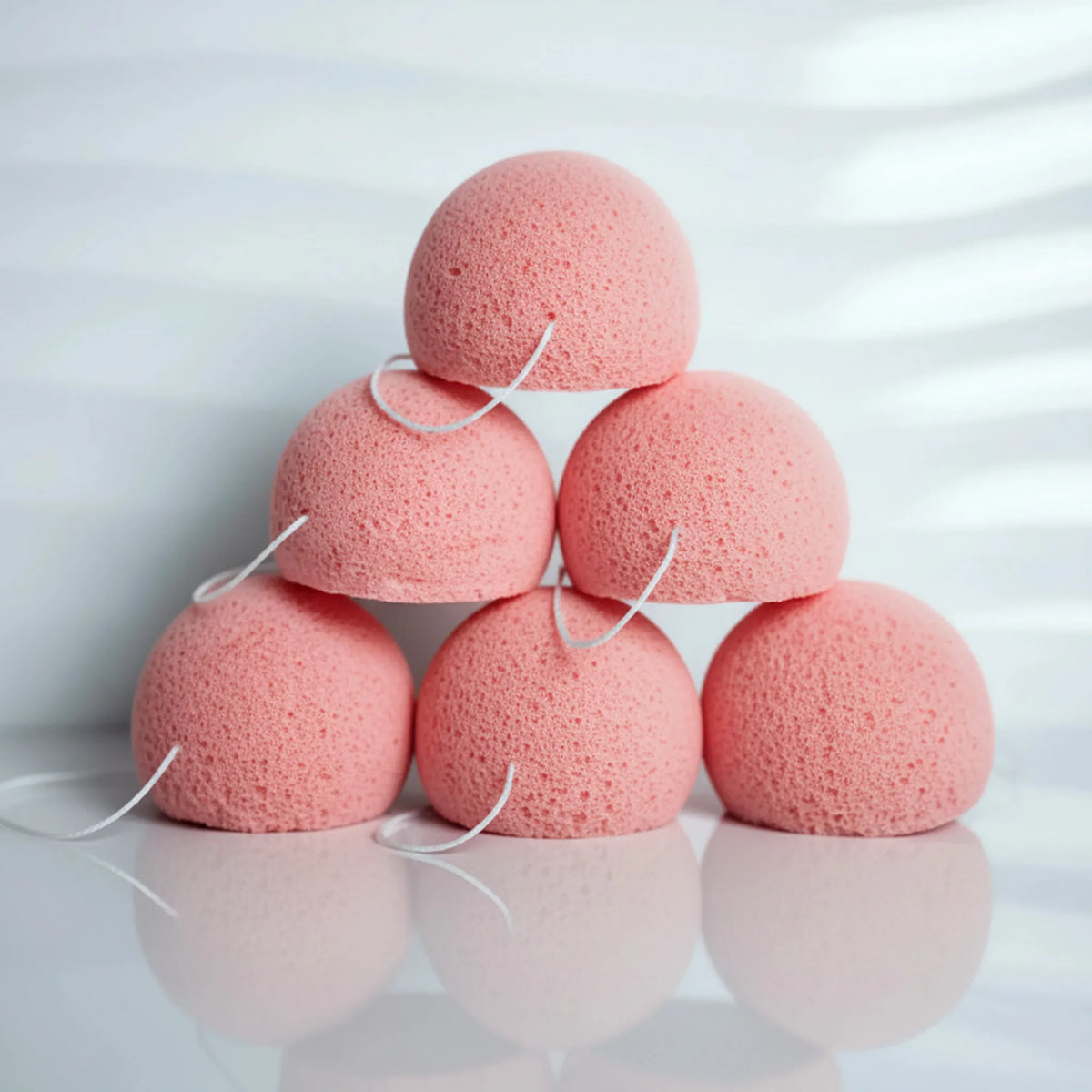 Six pink clay konjac sponges arranged in a pyramid stack on a reflective white surface with a soft white background.