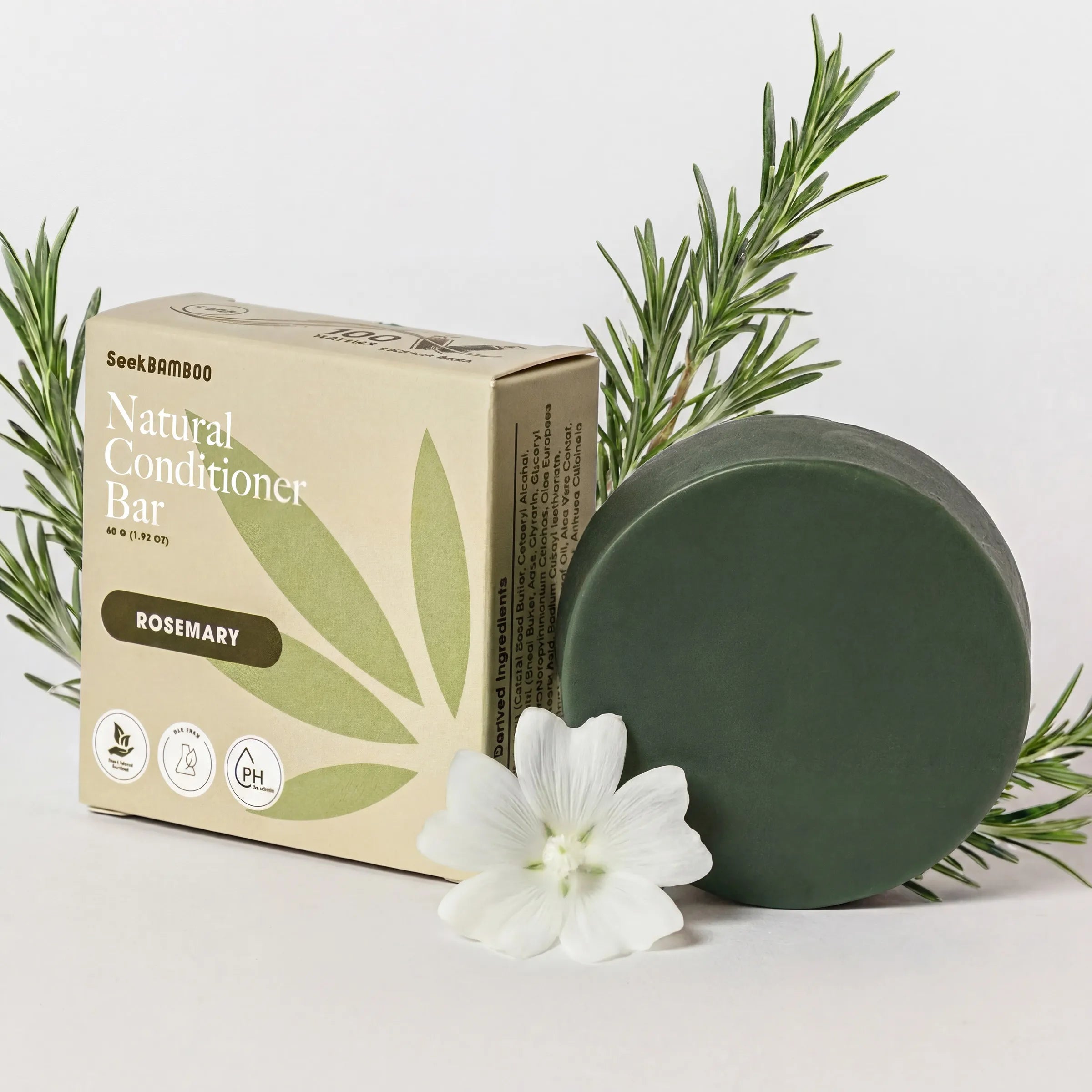 Natural gray hair conditioner bar in rosemary scent, featuring a dark green round bar of conditioner next to its beige cardboard box, styled with fresh rosemary sprigs and a white flower on a clean white background.
