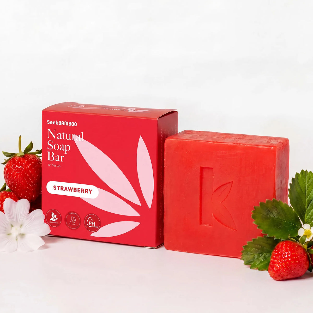 A strawberry soap bar next to its red packaging box, styled with fresh strawberries and green leaves on a white background.