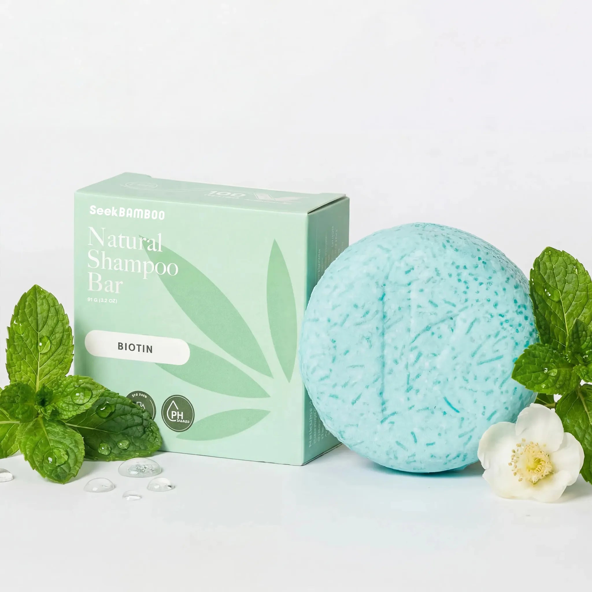 volume shampoo bar with biotin, featuring a light blue-green textured round shampoo bar next to its mint green packaging box, styled with fresh mint leaves, a white flower, and water droplets on a clean white background.